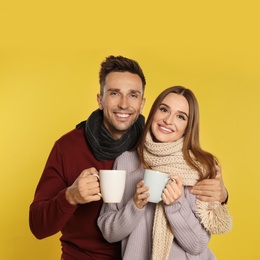 Photo of Happy young couple in warm clothes with cups on yellow background. Winter season