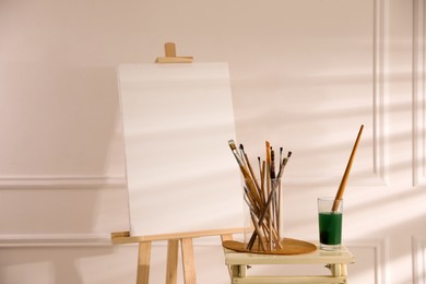 Photo of Wooden easel with empty canvas and brushes in art studio