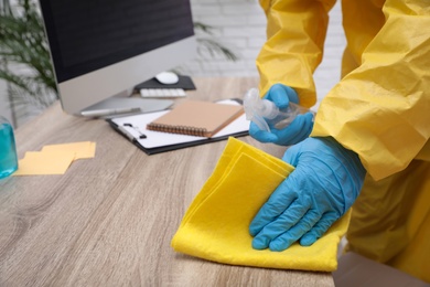 Photo of Janitor in protective suit disinfecting office furniture to prevent spreading of COVID-19, closeup