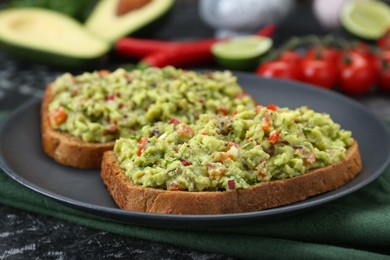 Photo of Slices of bread with tasty guacamole and ingredients on black textured table, closeup