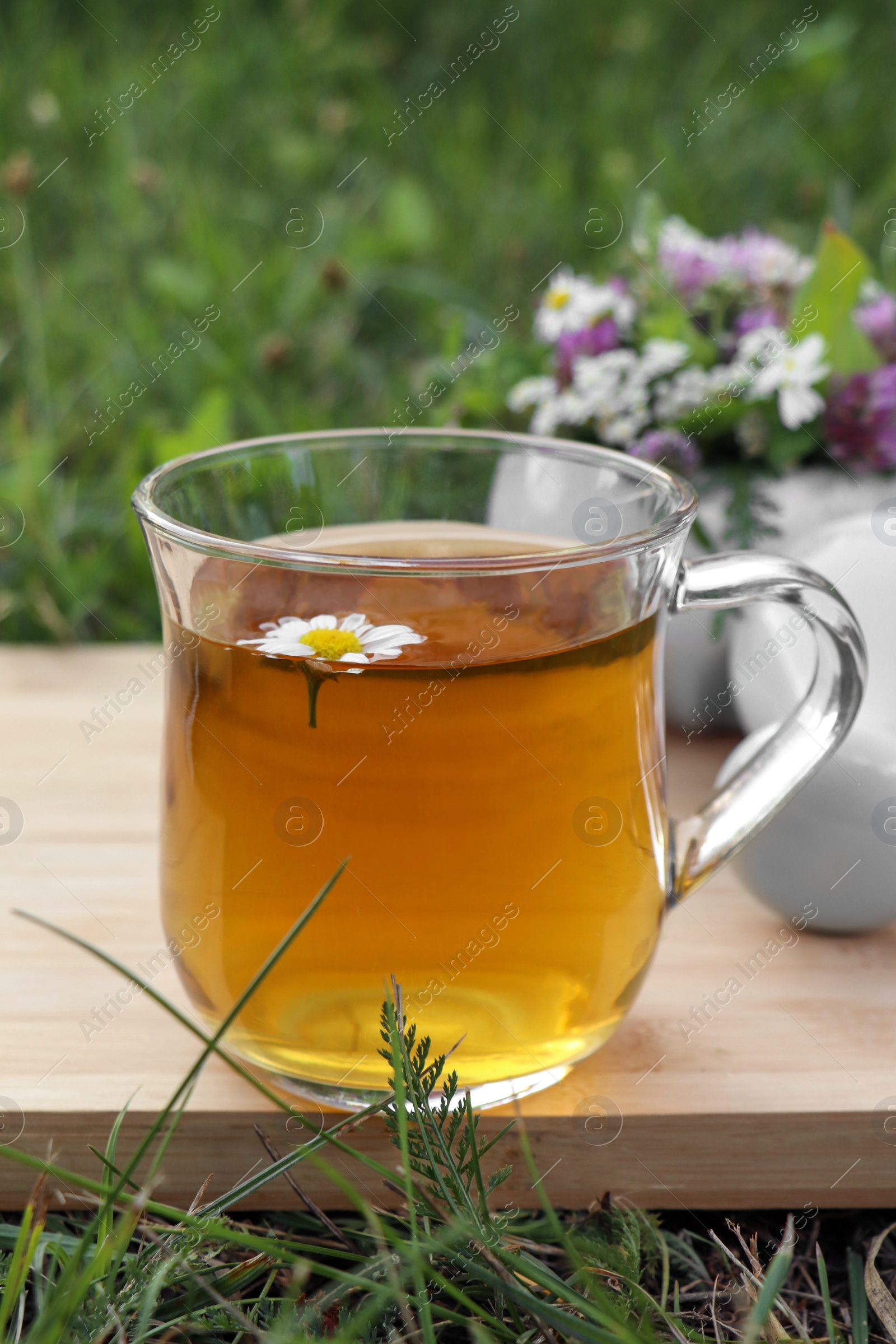 Photo of Cup of aromatic herbal tea, pestle and ceramic mortar with different wildflowers on green grass outdoors