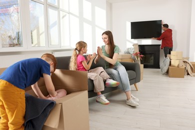Photo of Family settling into new home. Dad setting up TV, happy mom and daughter playing on couch, son unpacking his boxes. Moving day