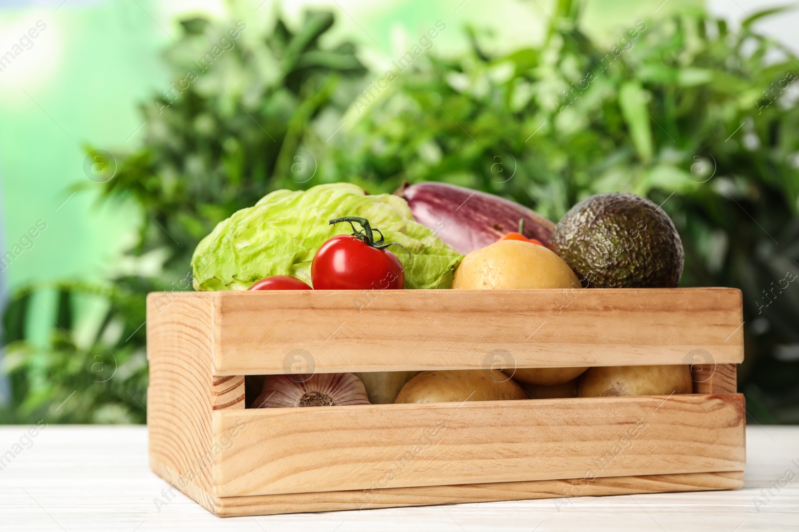 Photo of Fresh vegetables in crate on white wooden table against blurred green background