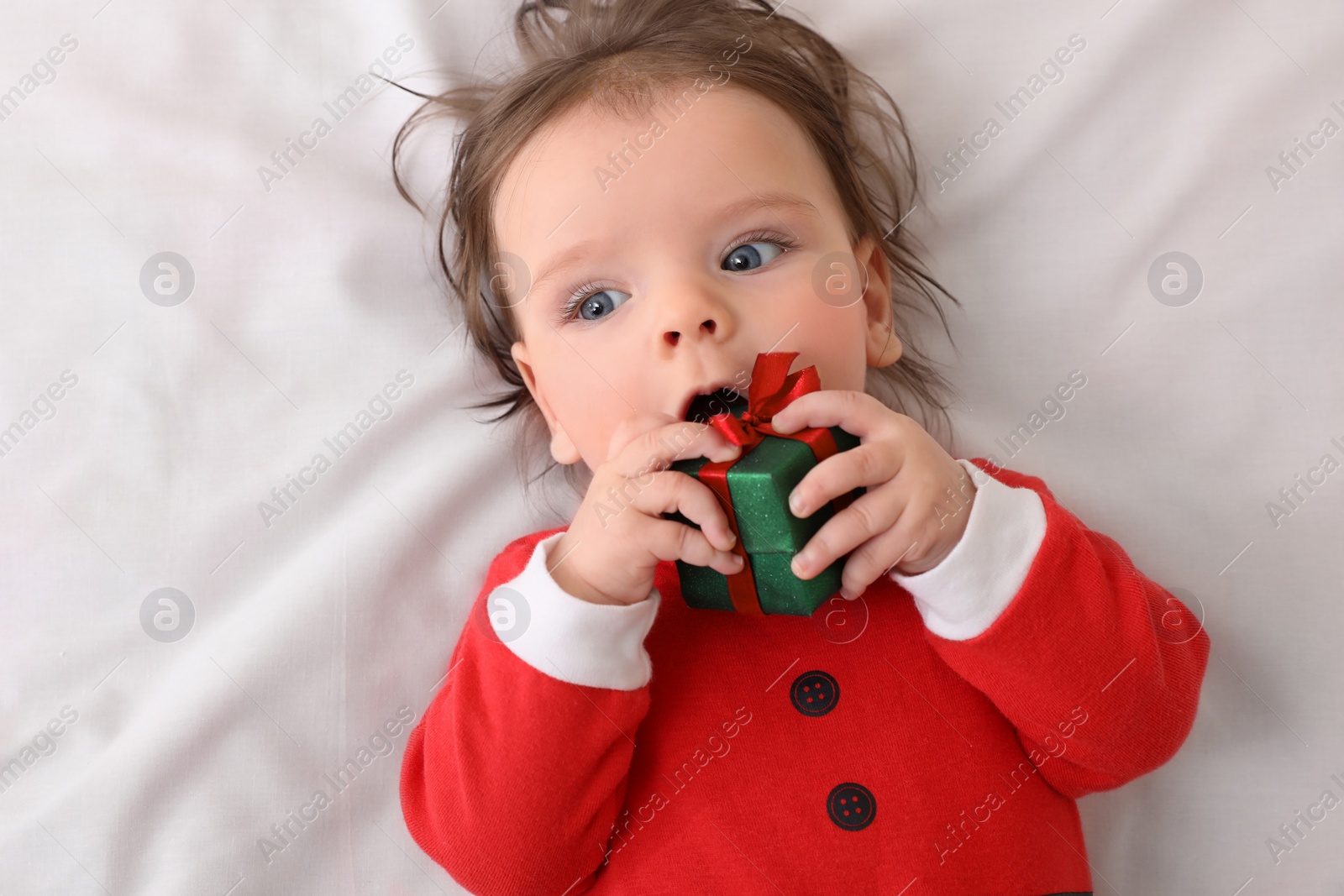 Photo of Cute baby wearing festive Christmas costume with gift box on white bedsheet, top view