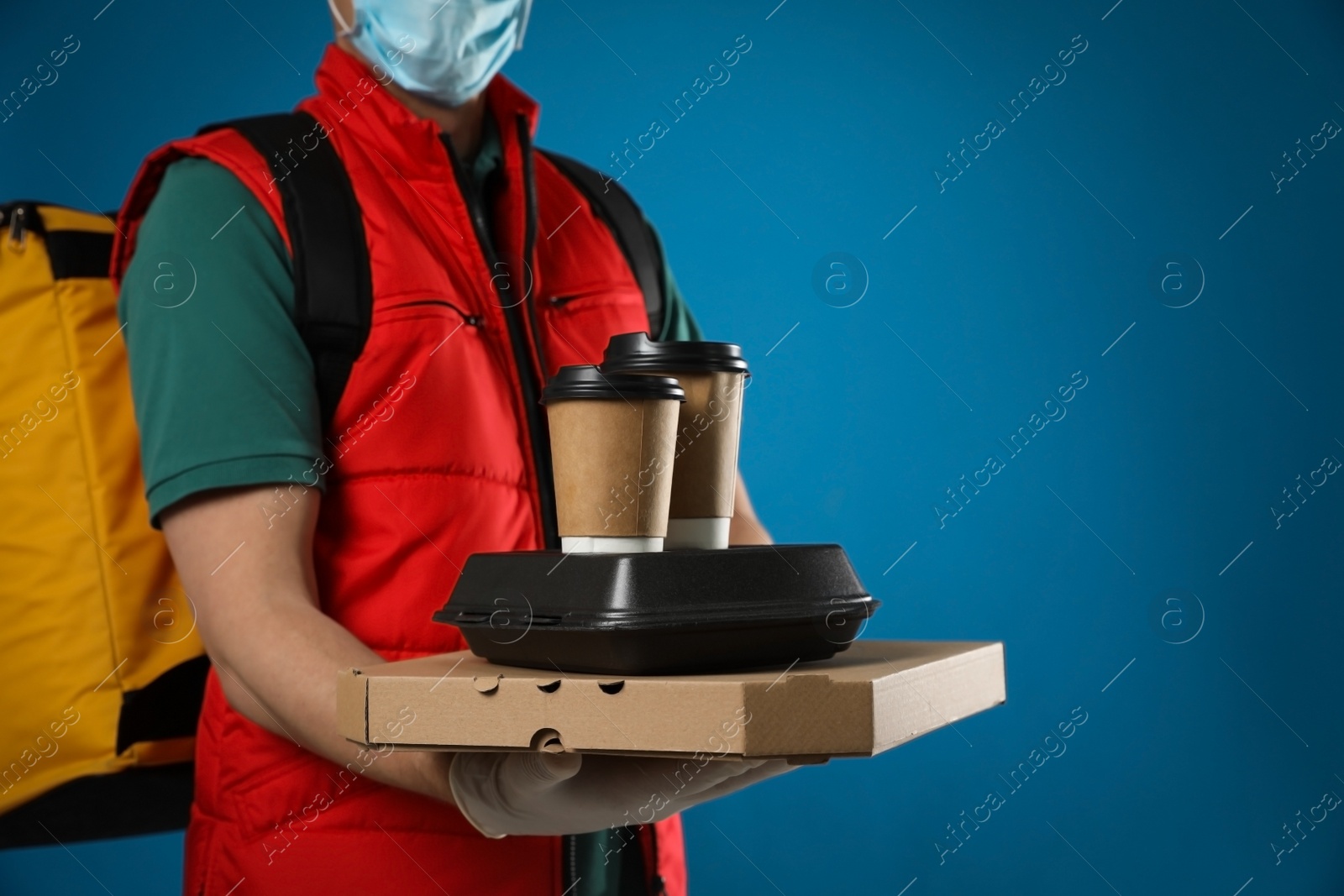 Photo of Courier in protective mask and gloves holding order on blue background, closeup. Food delivery service during coronavirus quarantine