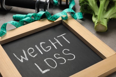 Photo of Small chalkboard with phrase Weight Loss, measuring tape and broccoli on grey table, closeup