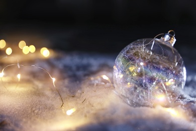 Photo of Christmas ball and fairy lights on white snow outdoors. Space for text