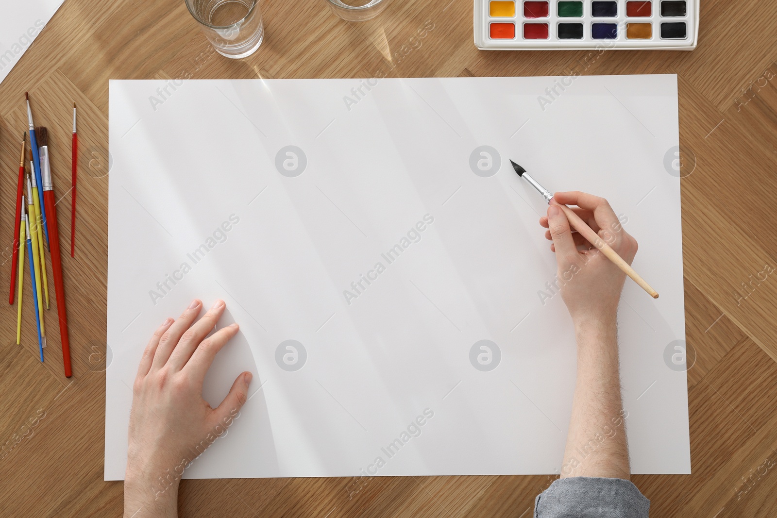 Photo of Man painting with watercolor on blank paper at wooden table, top view