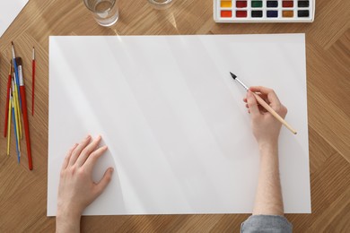 Photo of Man painting with watercolor on blank paper at wooden table, top view