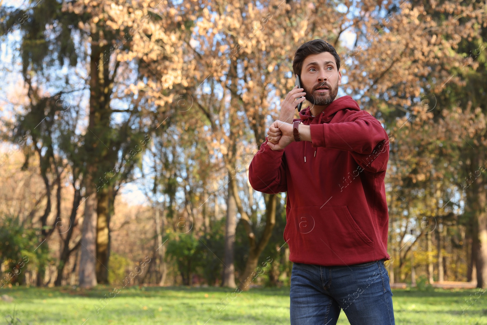 Photo of Emotional man talking by smartphone and checking time on watch in park, space for text. Being late concept