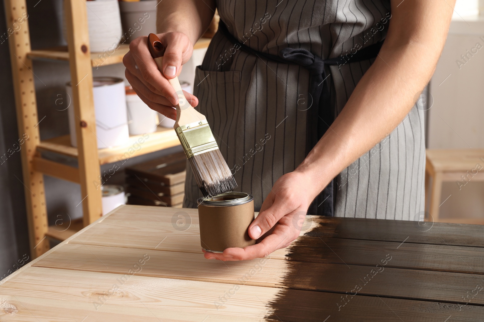 Photo of Man dipping brush into can of wood stain at wooden surface indoors, closeup