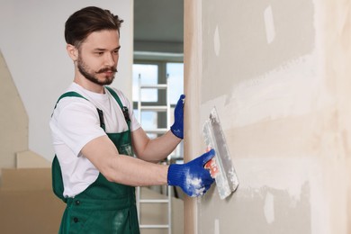 Worker in uniform plastering wall with putty knife indoors. Space for text