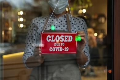 Photo of Woman in mask putting red sign with words Closed Due To Covid-19 hanging onto glass door, closeup