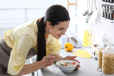 Photo of Young woman eating tasty vegetable soup at countertop in kitchen