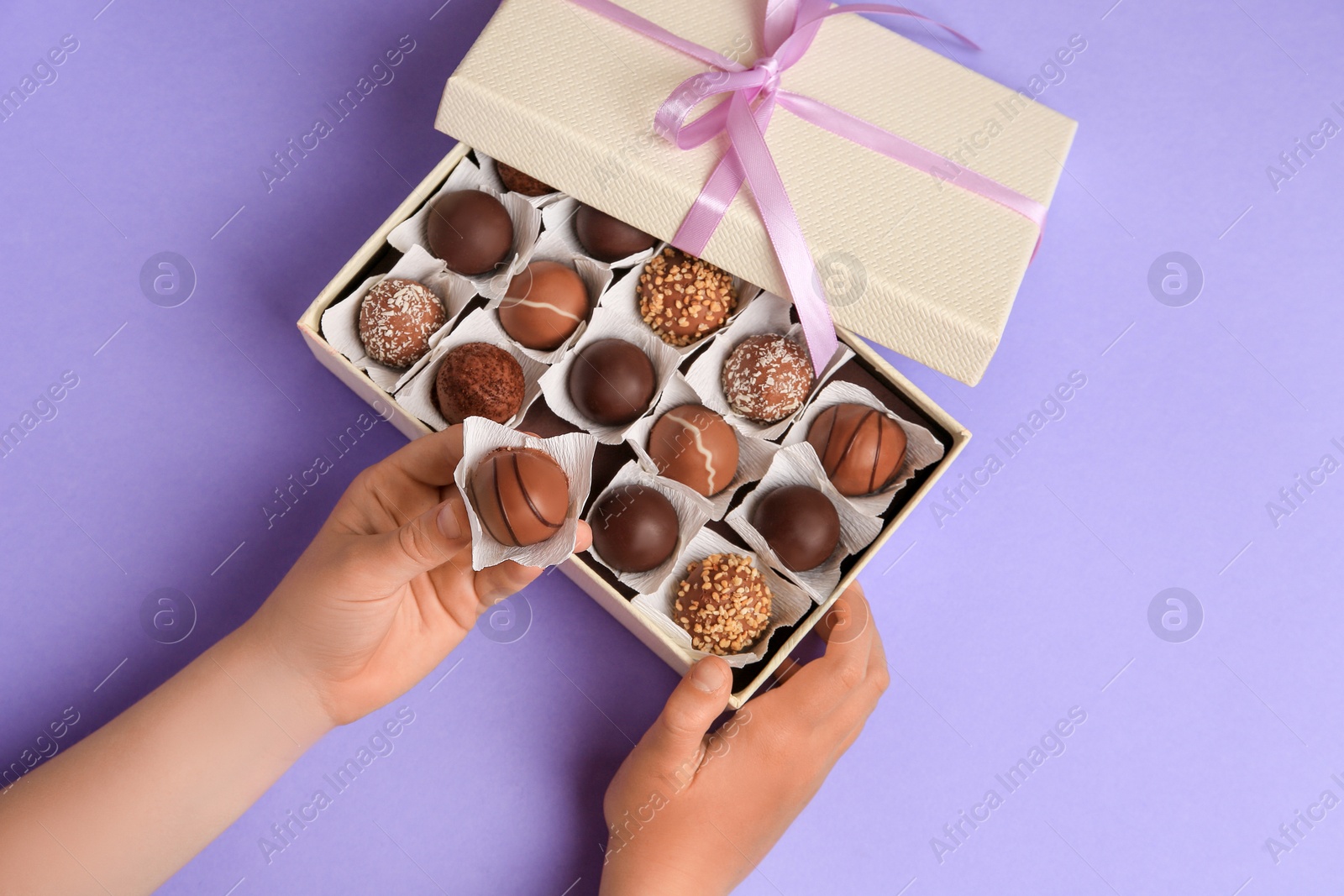 Photo of Child taking delicious chocolate candy from box on light purple background, top view