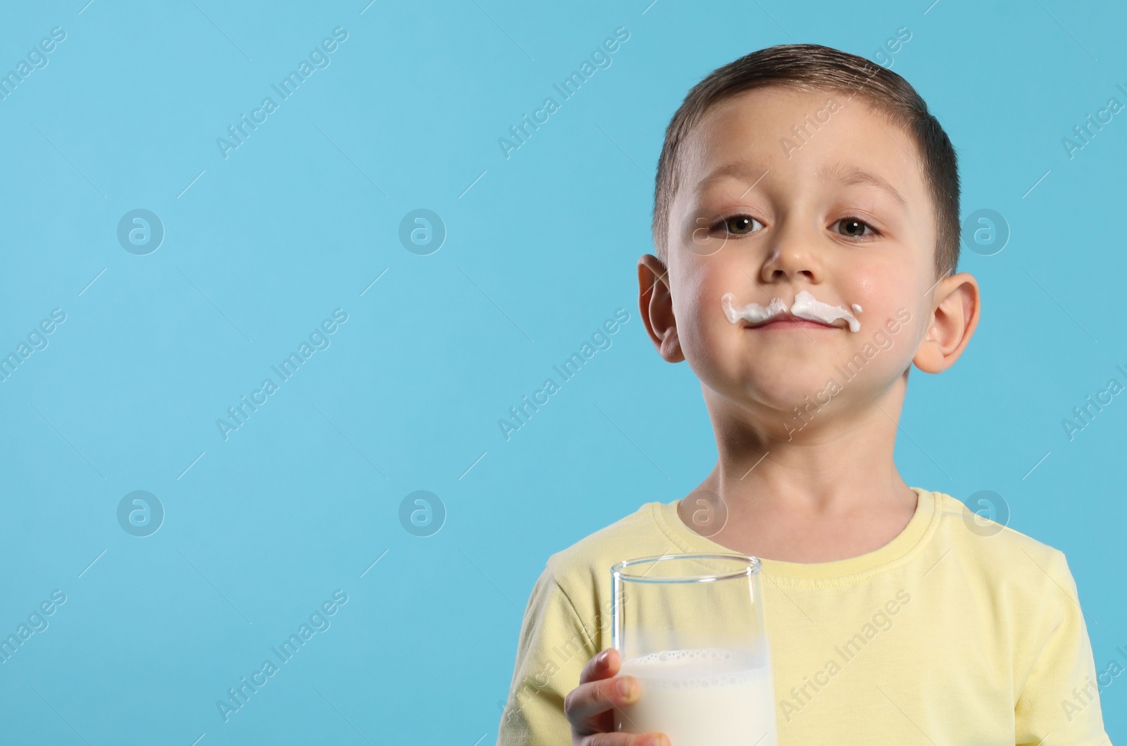 Photo of Cute boy with milk mustache holding glass of tasty dairy drink on light blue background, space for text