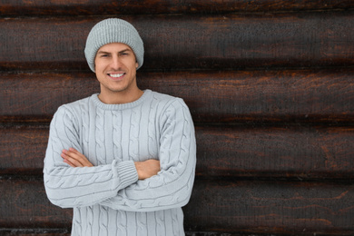 Photo of Handsome man wearing warm sweater and hat near wooden wall, space for text. Winter season