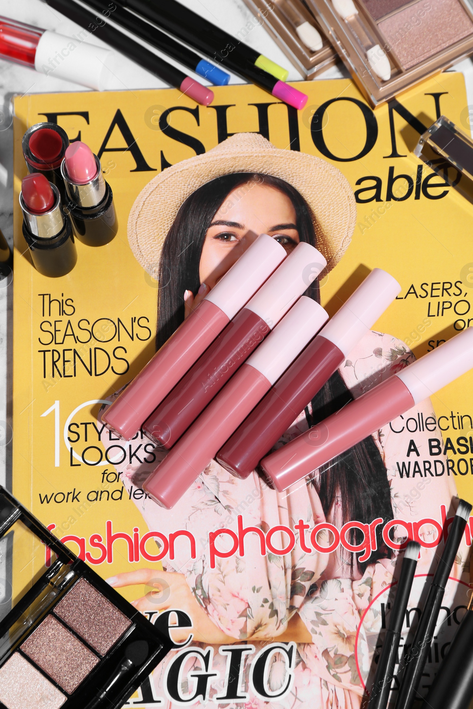 Photo of Bright lip glosses among different cosmetic products and fashion magazine on table, flat lay