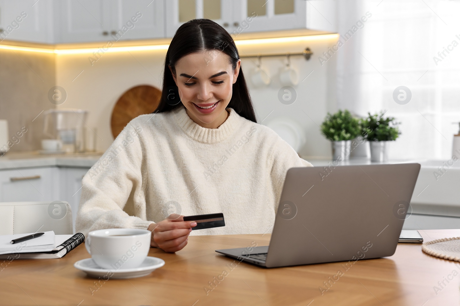 Photo of Happy young woman with credit card using laptop for shopping online at wooden table in kitchen