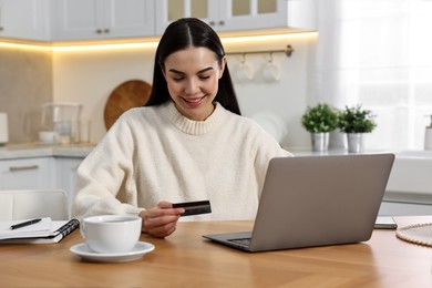 Photo of Happy young woman with credit card using laptop for shopping online at wooden table in kitchen