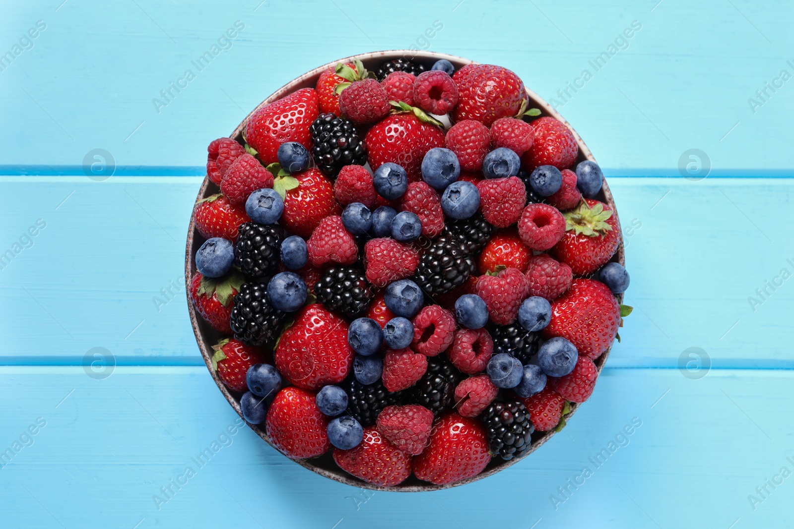 Photo of Different fresh ripe berries in bowl on light blue wooden table, top view