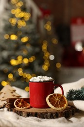 Cup of cocoa with marshmallows, cinnamon sticks, dry orange slices and Christmas decor on table against festive lights. Space for text