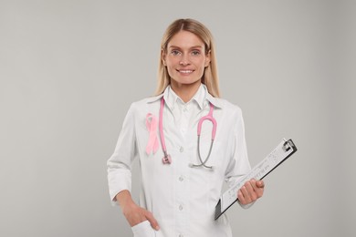 Photo of Doctor with pink ribbon, clipboard and stethoscope on light grey background. Breast cancer awareness