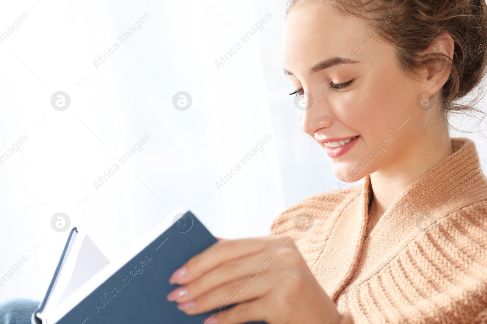 Photo of Portrait of beautiful teenage girl in warm cozy sweater reading book near window at home