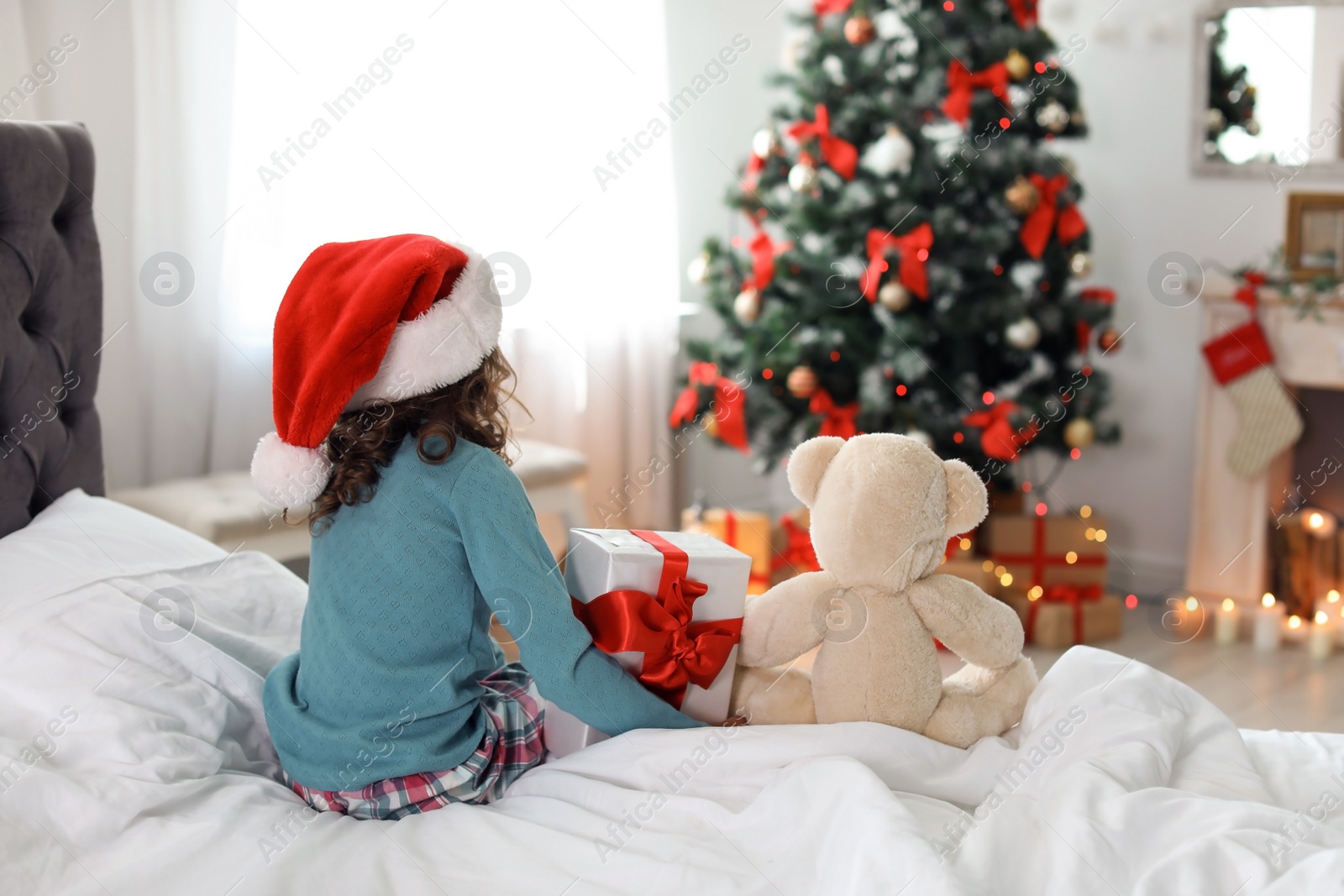 Photo of Cute little child in Santa hat with toy bear and Christmas gift box sitting on bed at home