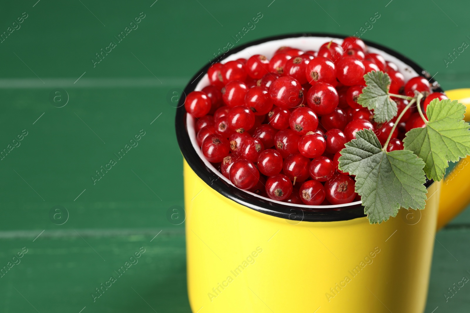 Photo of Ripe red currants and leaves in mug on green wooden table, closeup. Space for text