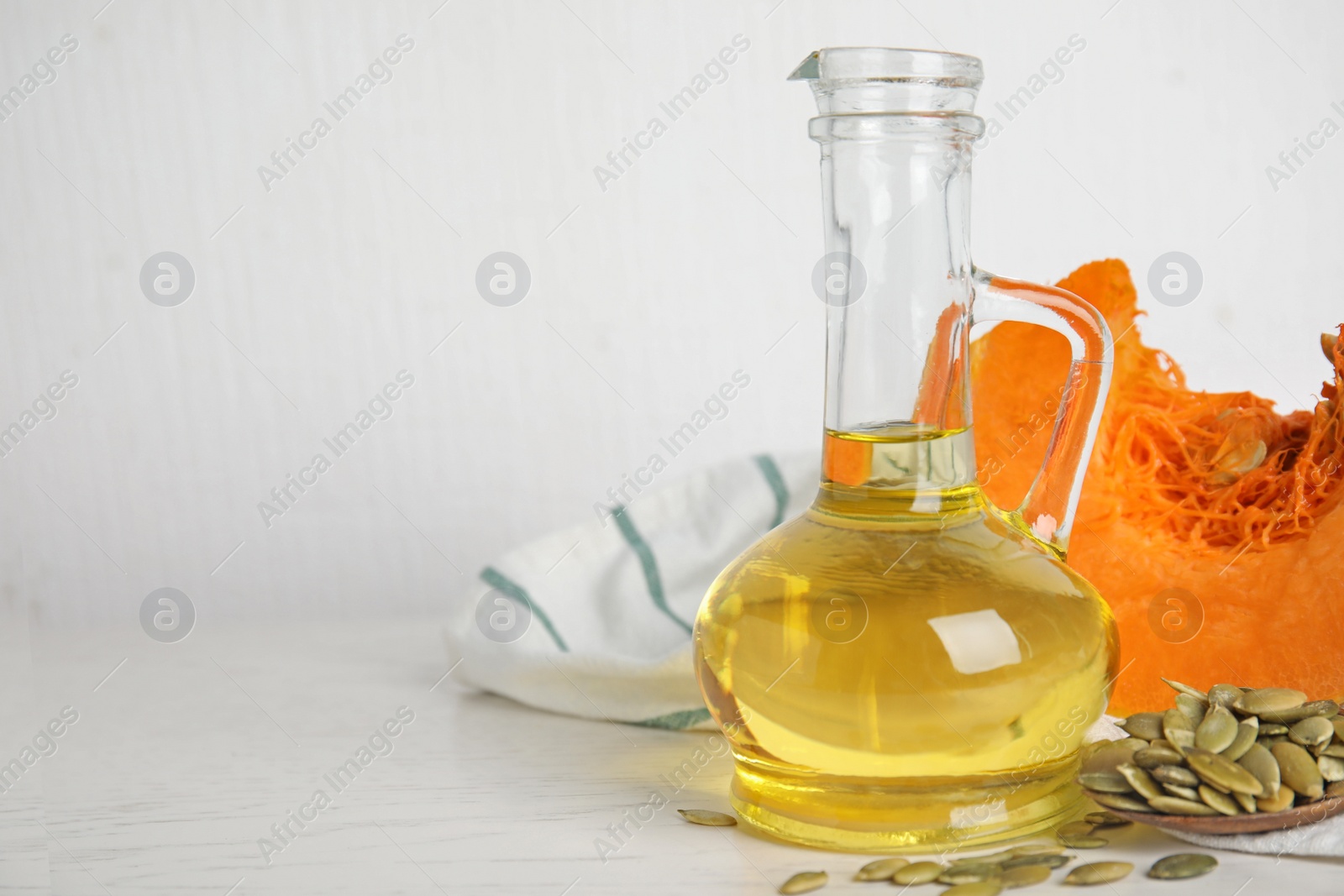 Photo of Glass jug of oil with cut pumpkin and seeds on white wooden table. Space for text