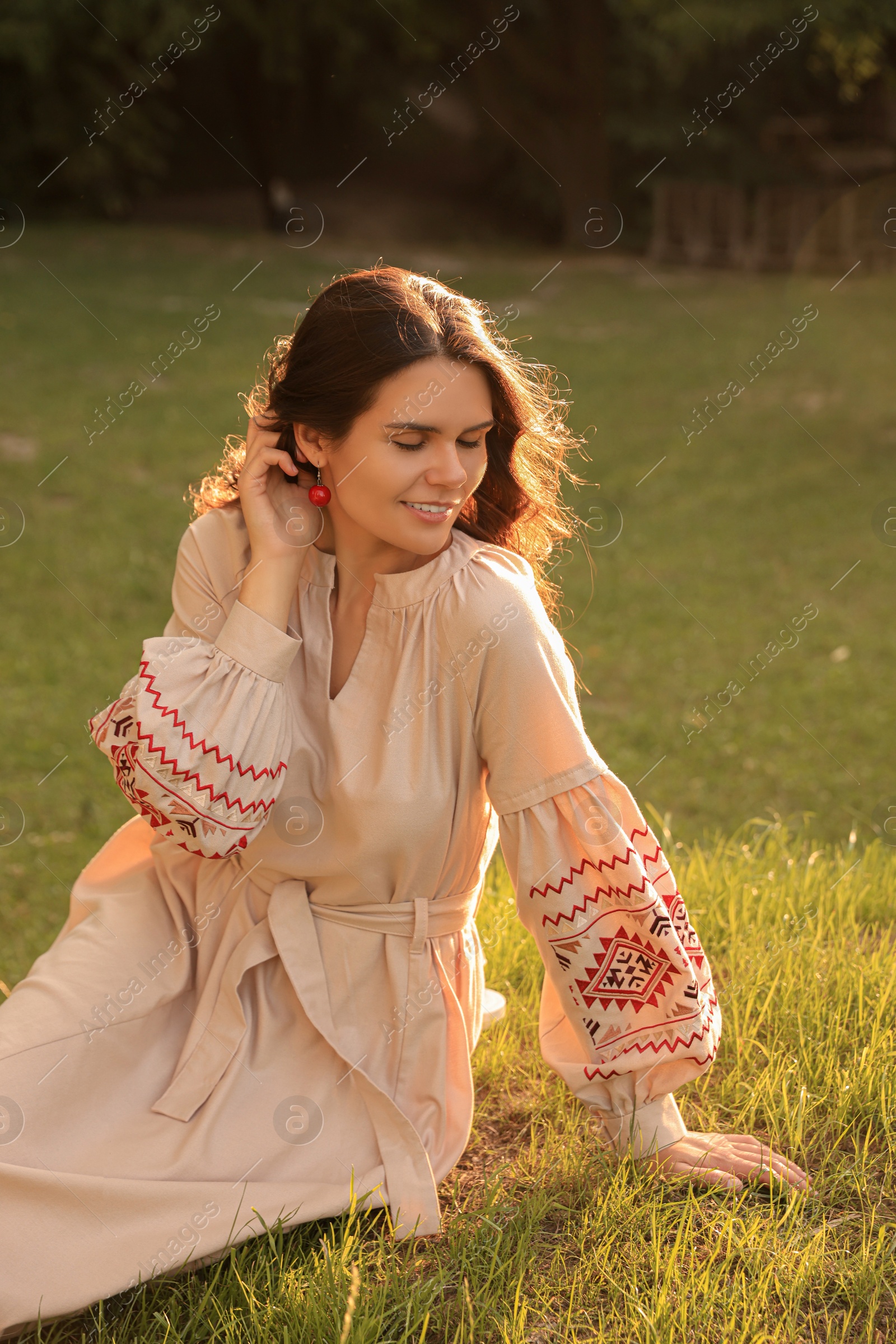 Photo of Beautiful woman in embroidered dress sitting on green grass outdoors. Ukrainian national clothes