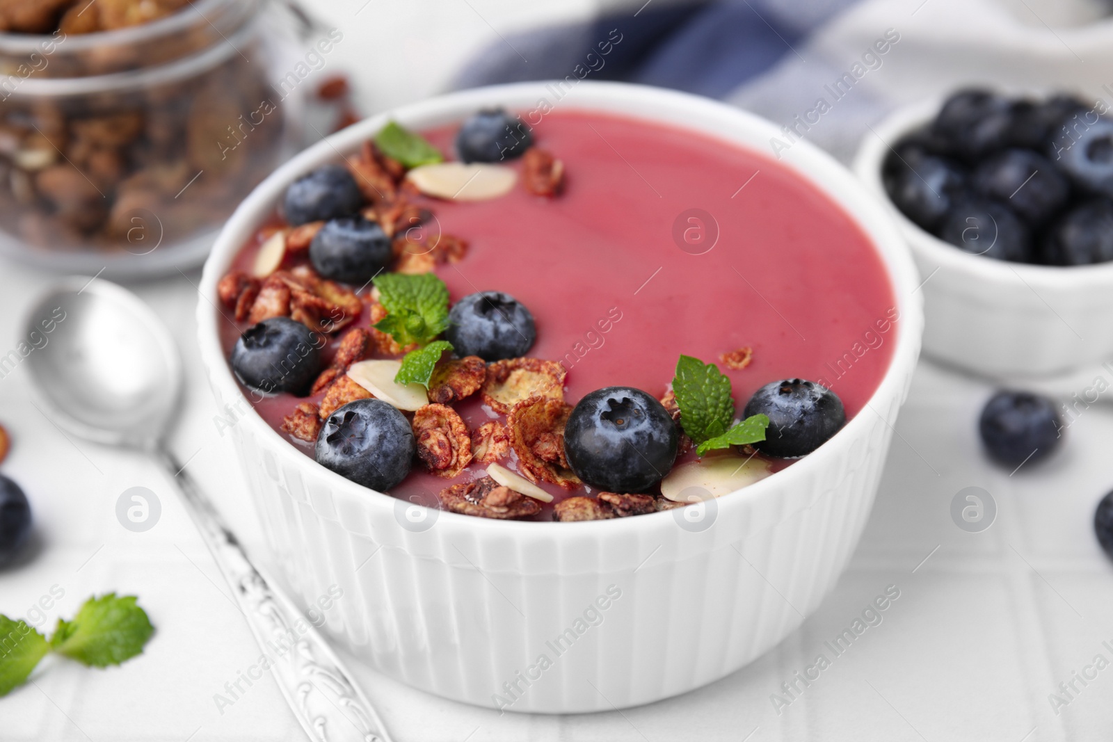 Photo of Bowl of delicious smoothie,served with fresh blueberries and granola on white tiled table, closeup