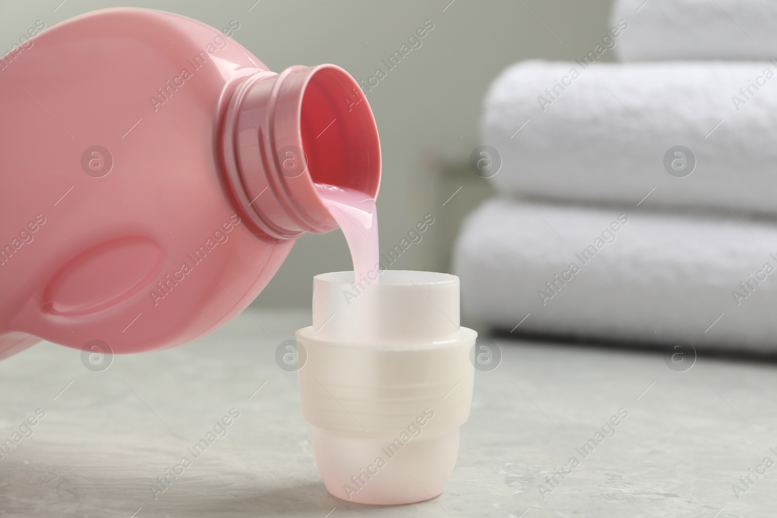 Photo of Pouring laundry detergent from bottle into cap on light grey marble table, closeup