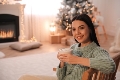 Young woman with cup of hot drink at home, space for text. Christmas celebration