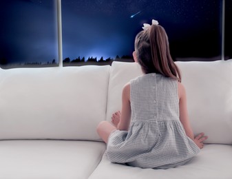 Cute little girl sitting on sofa near window and looking at shooting star in beautiful night sky