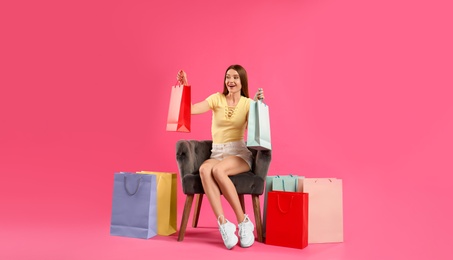 Beautiful young woman with paper shopping bags in armchair on pink background