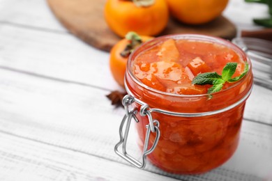 Jar of tasty persimmon jam and ingredients on white wooden table. Space for text