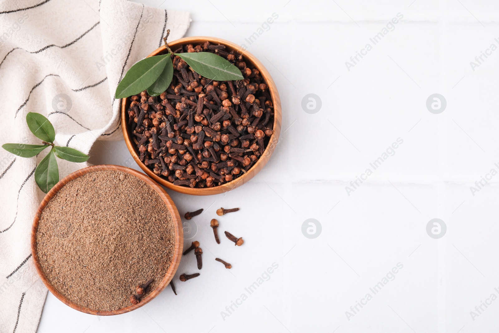 Photo of Aromatic clove powder and dried buds in bowls on white tiled table, flat lay. Space for text
