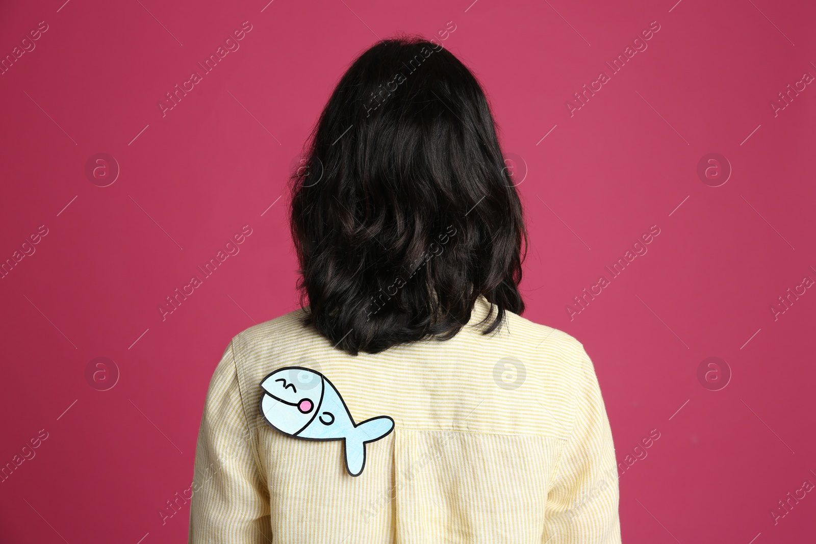 Photo of Woman with paper fish on back against pink background. April fool's day