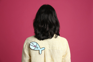 Photo of Woman with paper fish on back against pink background. April fool's day