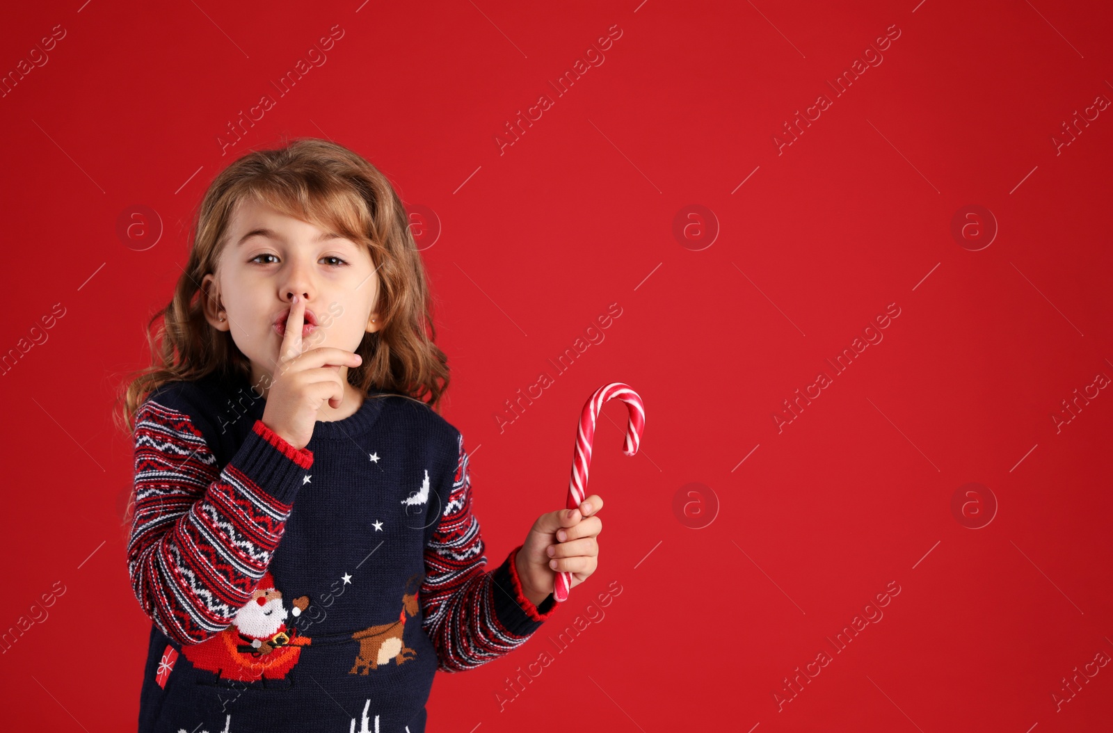 Photo of Cute little girl with sweet Christmas candy cane showing silence gesture against red background