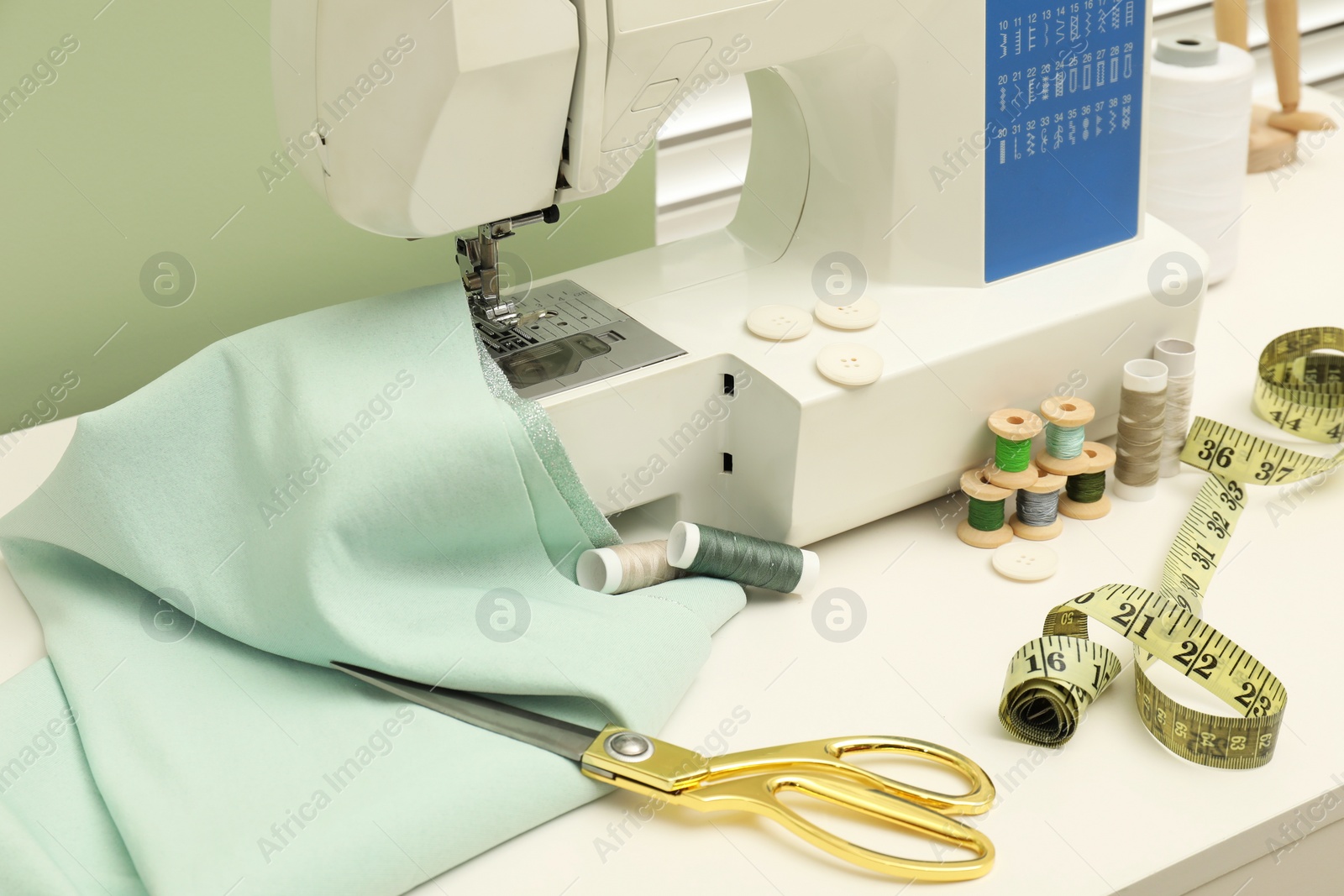 Photo of Sewing machine with fabric, scissors, measuring tape and threads on white table, closeup