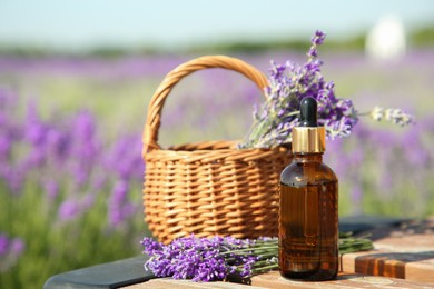 Photo of Bottle of essential oil and wicker bag with lavender flowers on wooden table in field outdoors, space for text