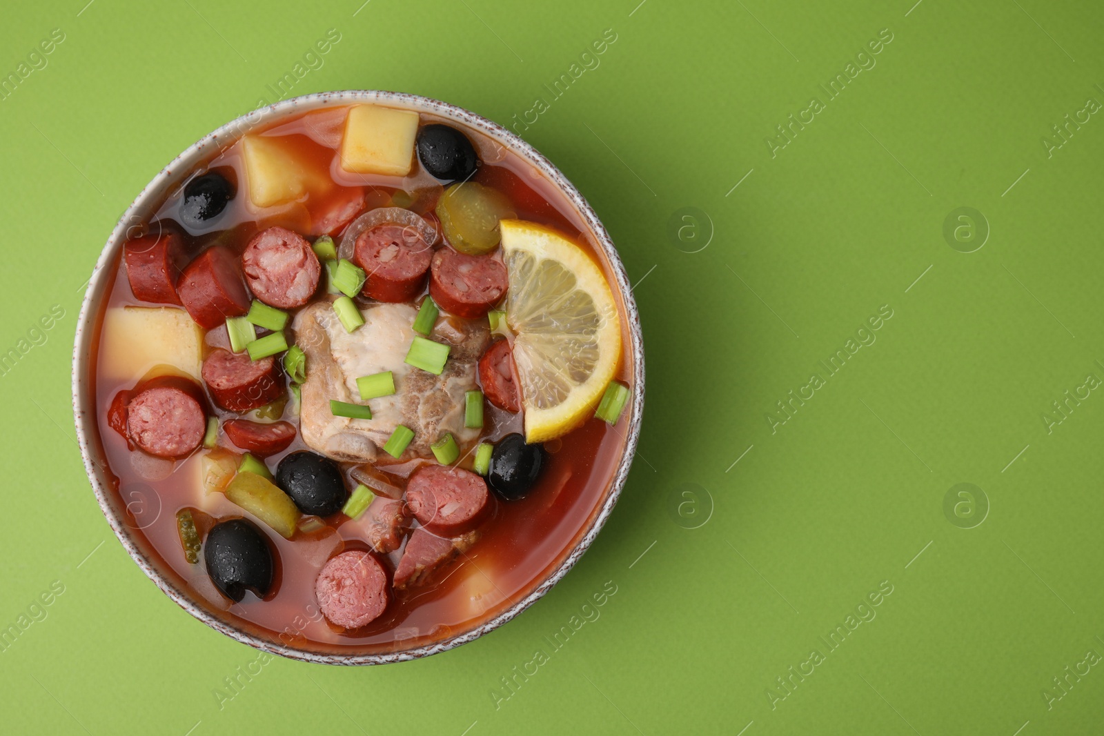 Photo of Meat solyanka soup with thin dry smoked sausages in bowl on light green background, top view. Space for text