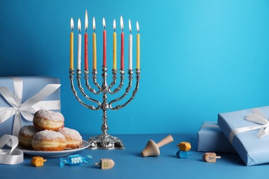 Photo of Hanukkah celebration. Menorah with burning candles, dreidels, donuts and gift boxes on light blue table, space for text