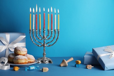 Hanukkah celebration. Menorah with burning candles, dreidels, donuts and gift boxes on light blue table, space for text