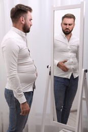 Upset man wearing tight shirt in front of mirror at home. Overweight problem