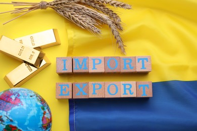 Words Import and Export made of wooden cubes, ears of wheat near globe and Ukrainian flag on yellow background, flat lay