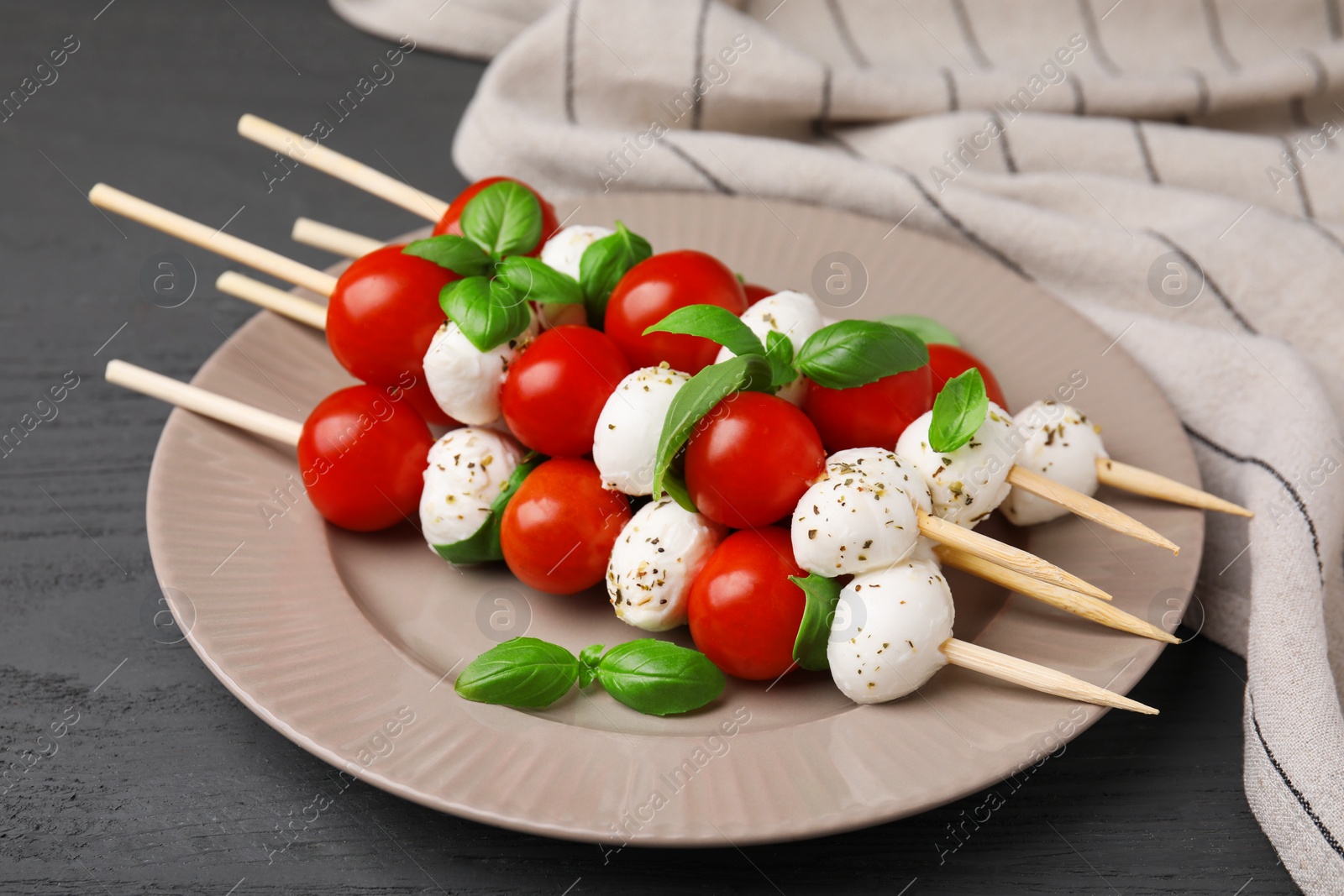 Photo of Caprese skewers with tomatoes, mozzarella balls, basil and spices on grey wooden table, closeup